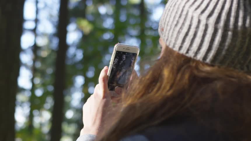 Woman who wears wooly hat takes picture of trees with smartphone camera in the forest by day