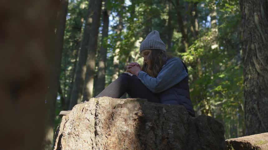 Woman wearing wooly hat sits on tree trunk and clasps hand in the forest by day
