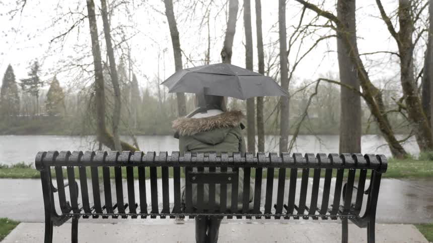 A woman sits on a park bench facing a river while holding an umbrella in the snow