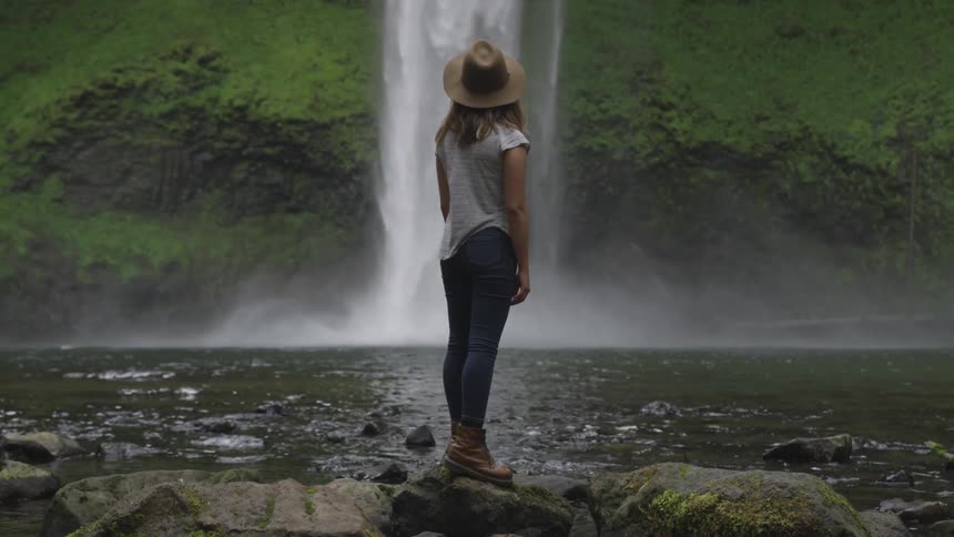 White woman standing at rocks near a lake and watching a waterfall at a distance