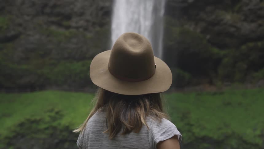 Woman wearing hat stands watching waterfall