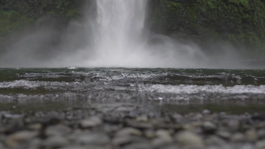 A waterfall creates powerful waves in a stream toward a shore of pebbles