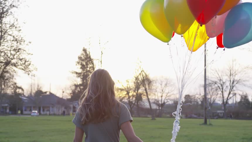 Blonde woman walks in the grass field and holds a bunch of colorful helium balloons at dusk