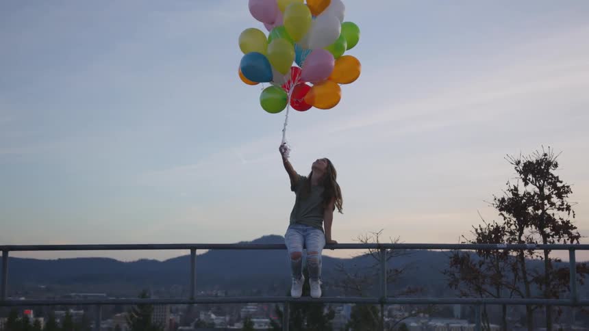 Woman holding bunch of balloons sits on handrail with city in background