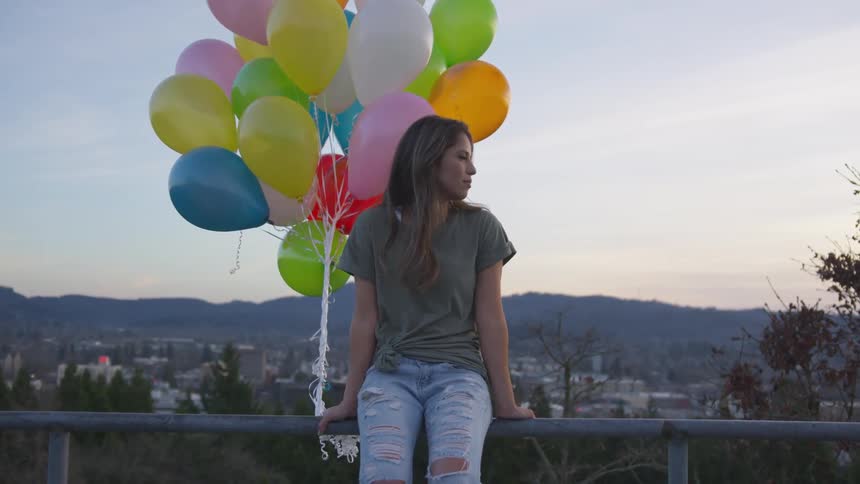 Woman sitting on a railing holding a bunch of colorful balloons