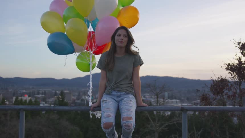 Woman sitting on metal bar while holding several balloons and contemplating the view