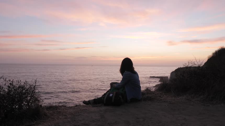 A female is walking and sits down facing the ocean