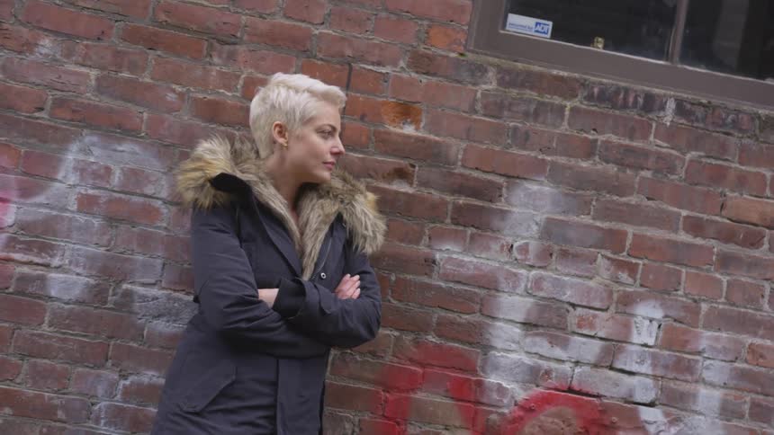 A woman in a blue coat standing against a graffiti covered brick wall