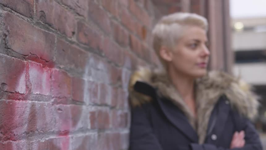 A woman wearing a parka leans against a brick wall in an alley