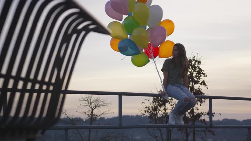 Teenage girl sits on a rail holding a lot of colored balloons