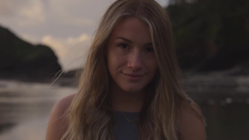 A blonde woman standing in front of a lake stares at the camera