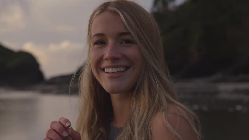 A young woman smiles and laughs while touching her hair and starting to turn near the water