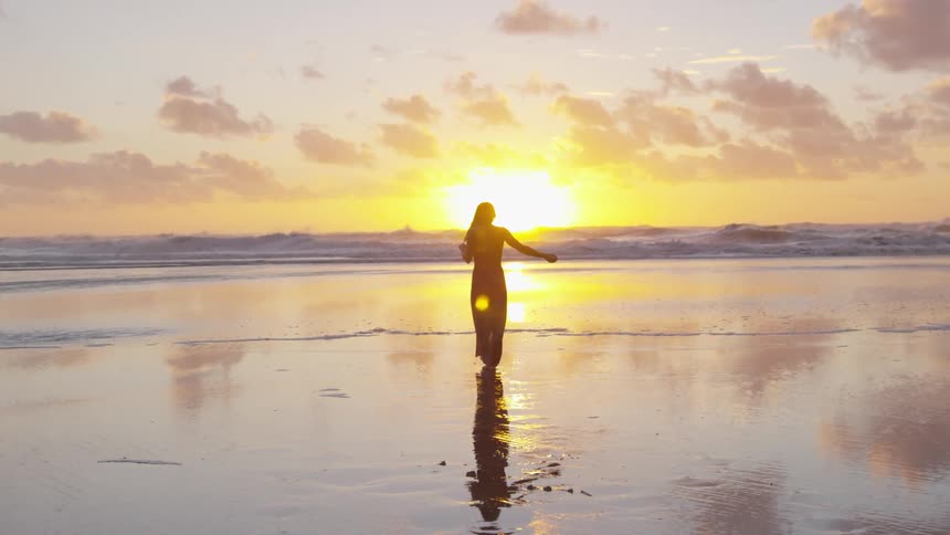 Woman combs hair as she walks across beach sand toward sun rising over ocean