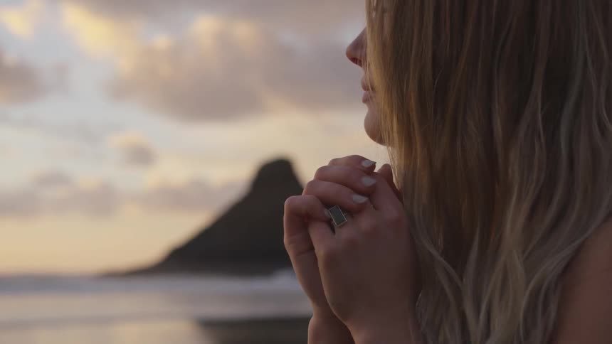 Blonde woman clasping her hands looks at sunset on the beach next to the ocean at dusk