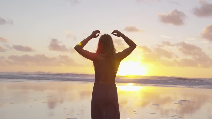 A woman lifts her arms into the air on a sandy beach at sunset