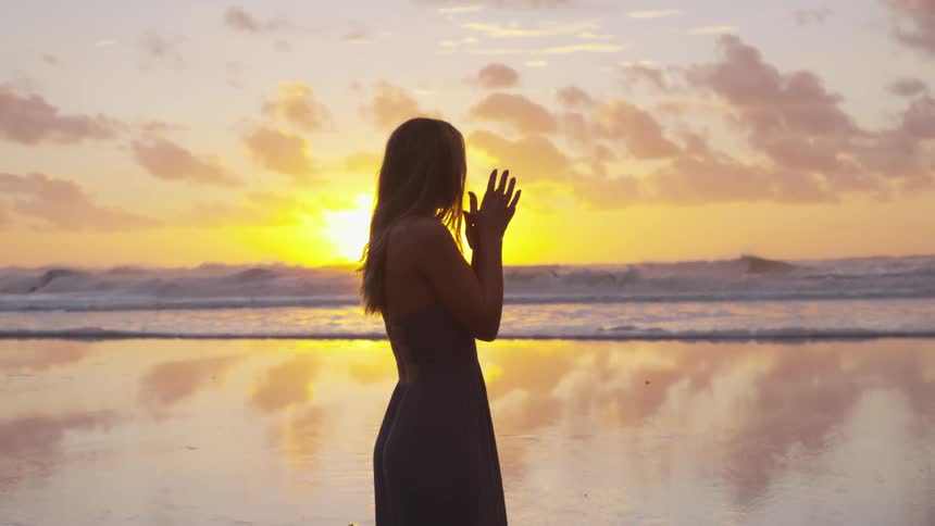 Woman clasps hand on the reflective sand beach at sunset next to the sea