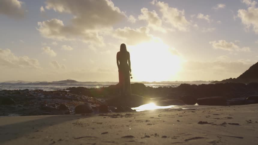 Woman standing at the beach holding a flower and looking at the horizon