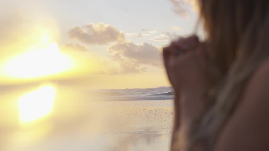 Blonde woman clasp hands on the sand beach reflecting sunlight at sunset