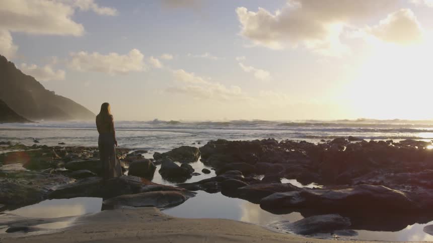 Woman stands on rocky beach at sunset