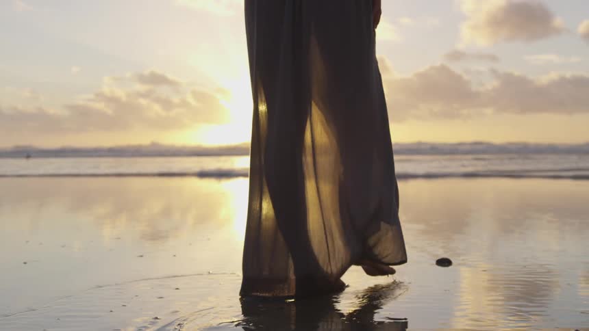 She's barefoot and walking on the beach needing footsteps wearing a gown at sunset