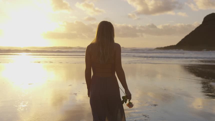 A woman carrying a rose walks and starts to twirl on the beach at sunset