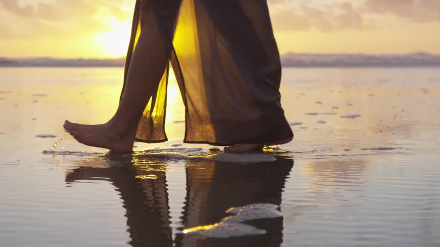 Woman walks barefoot in filmy dress across wet sand on beach