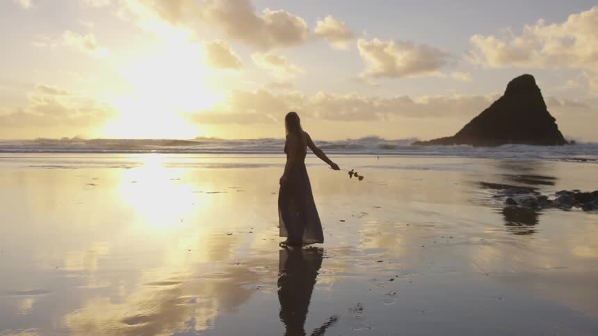 A woman holding a rose turns in circles on a beach