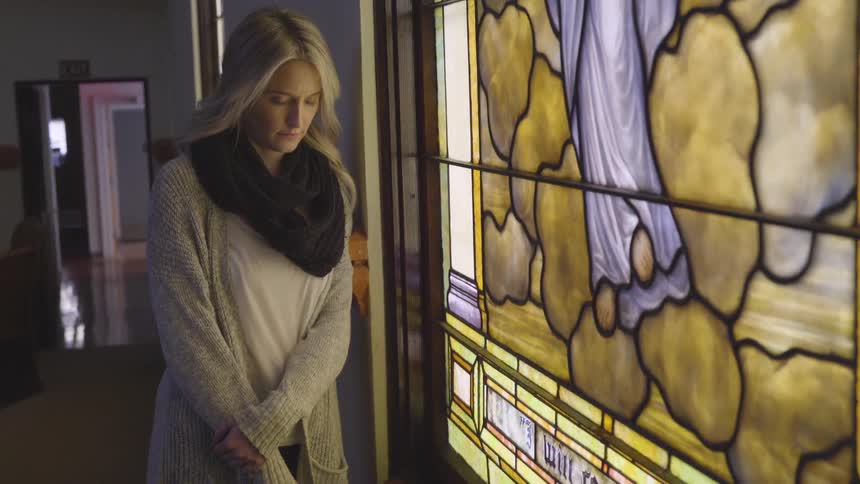 A young woman with blonde hair stands by a stained glass window