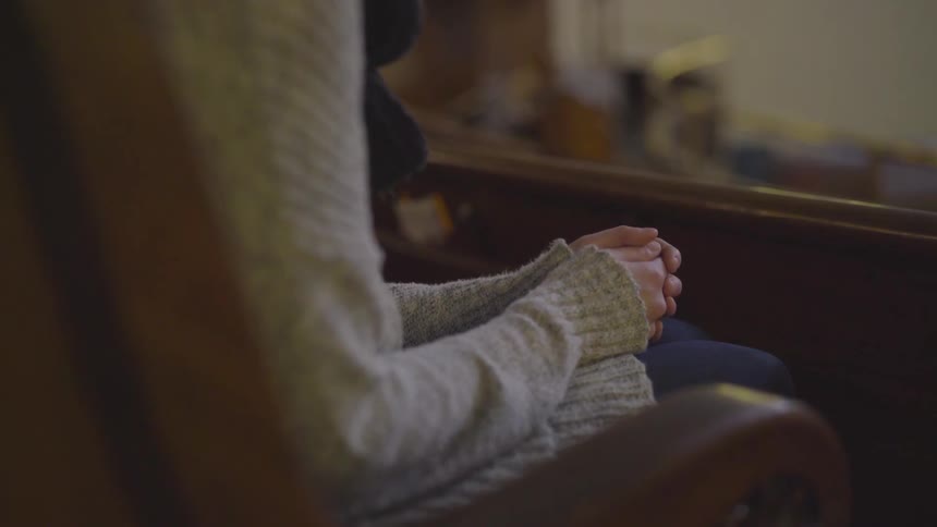 A woman wearing a long sleeved sweater sitting down clasps her hands in her lap