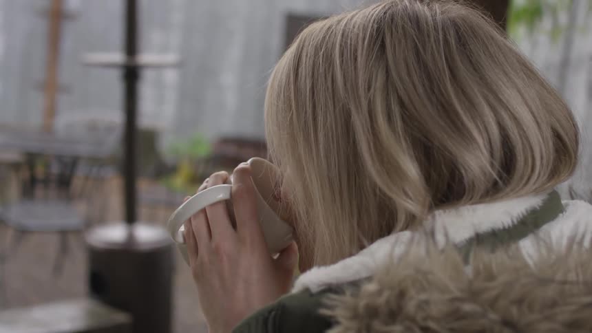 A seated blonde haired woman sipping coffee outside
