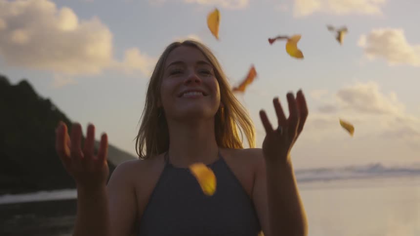 A woman laughs as she throws yellow flower petals in the air while standing on the beach