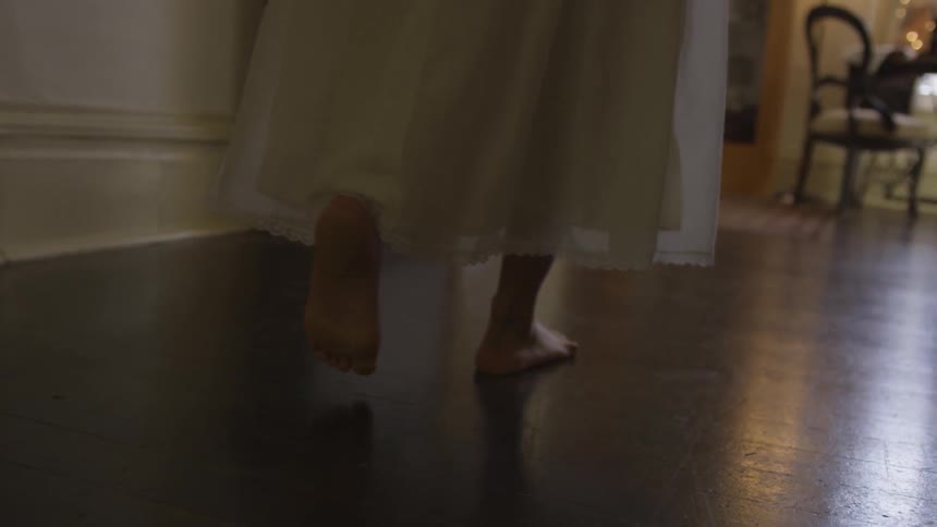 A woman in a white sleeping gown walking barefoot through a house