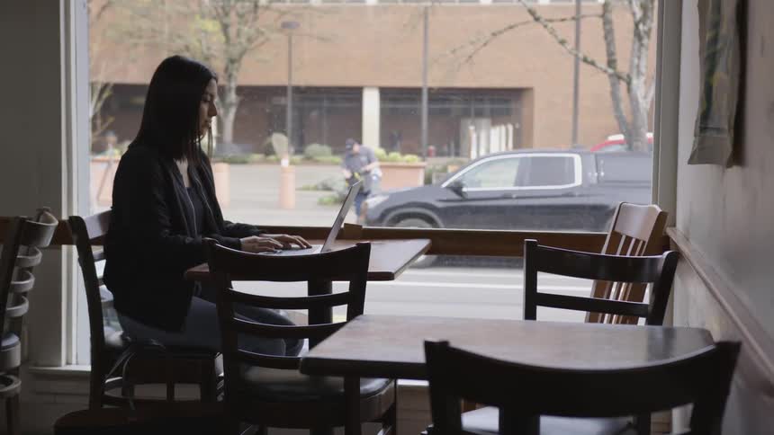 Woman sits at a table working on a laptop as a bicyclist rides by on the street