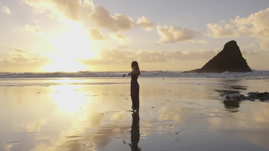 A woman in a long sheer skirt dances and twirls on the wet beach sand at sunset