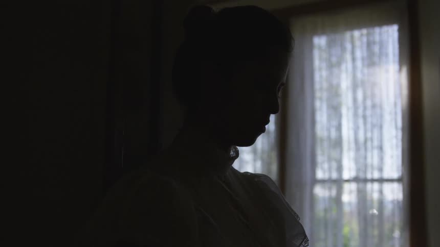 A woman wearing a vintage dress with a lace mock neck leans against a doorway with head bowed and eyes closed