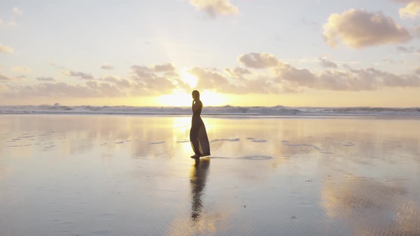 Woman walks on the sand reflecting sunset on beach next to the sea