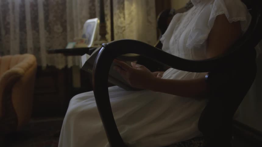Woman who wears white dress sits on chair and reads book in a dim room at night