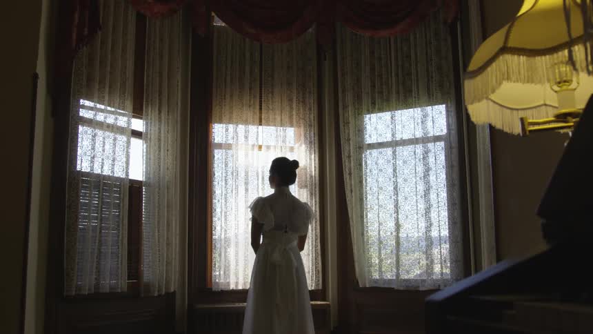 Woman stands in front of the windows and looks out through an opening in the curtains