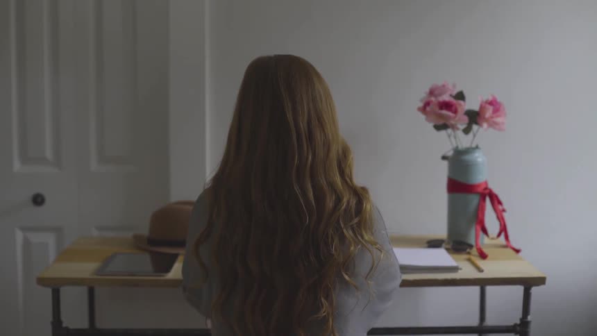 A female with long red hair cascading down her back sits at a small modern desk where she is looking over a notebook and a smart tablet