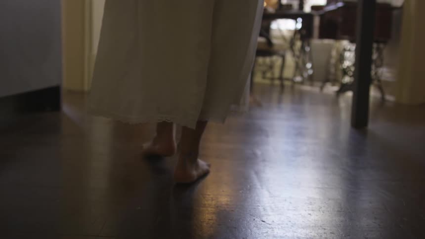 Woman walks barefoot down hall in white dress