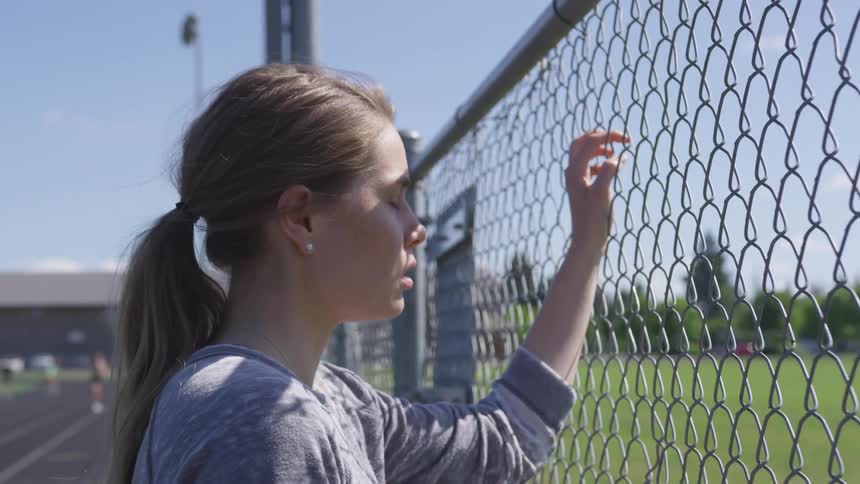 A teenage blonde white girl gets upset and stares through a chain link fence