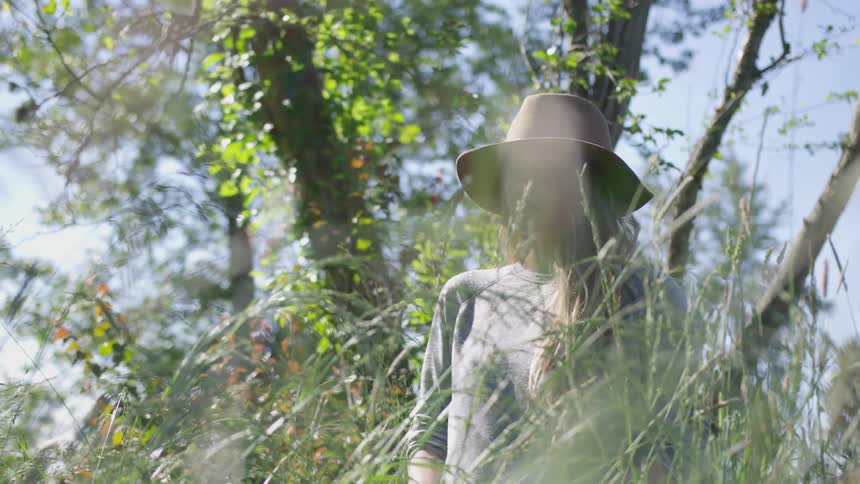 Young woman in a hat stands in a grass and looks into the camera