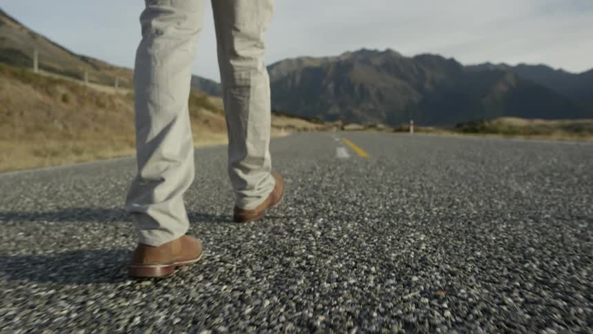 View of a man's legs and feet as he walks down a road towards mountains