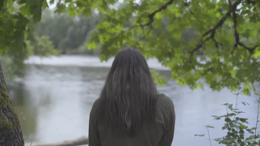 A woman stands beneath a tree on a riverbed