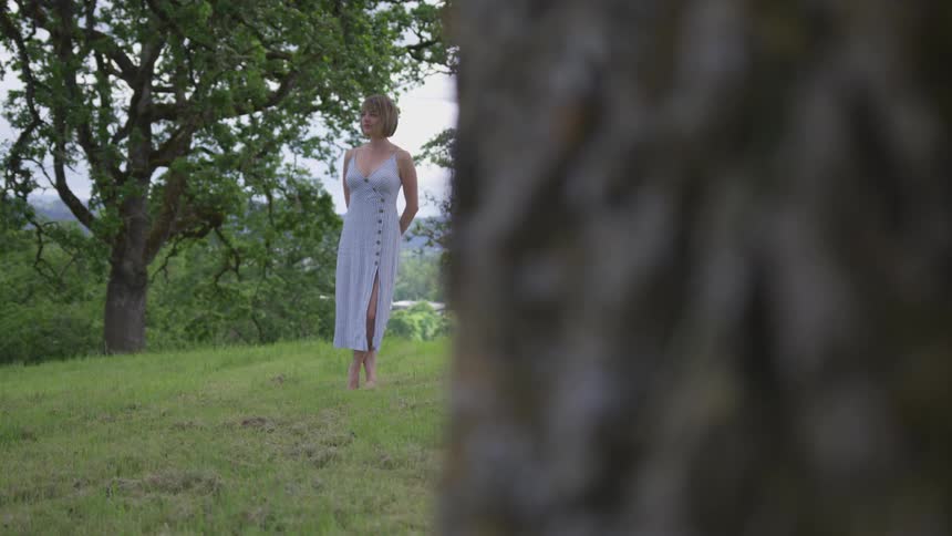 A woman in a white dress stands under trees in a grassy field