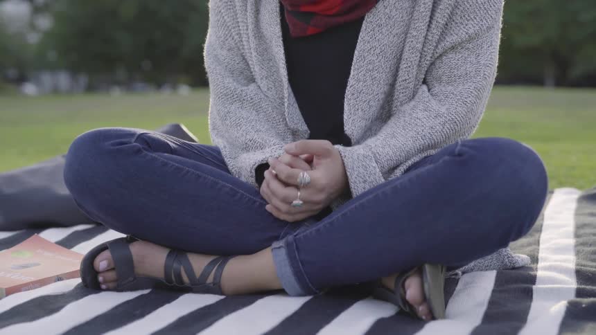 A woman sits on a blanket in a public park while clasping her hands