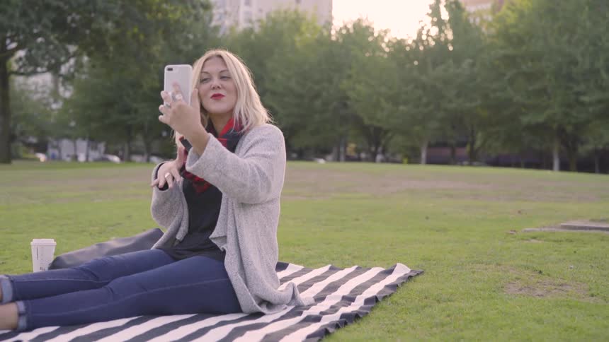 White woman talking to her cell phone while sitting above a towel in the park