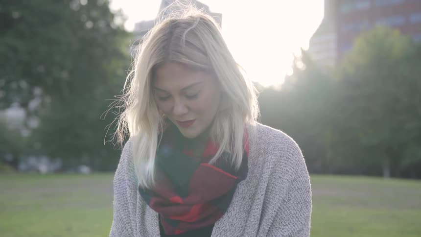 A blond woman wearing a sweater and scarf standing in a park with trees traffic and buildings behind her