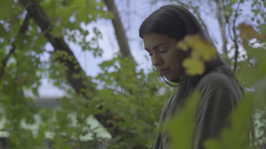 Woman stands among trees with head bowed and eyes closed