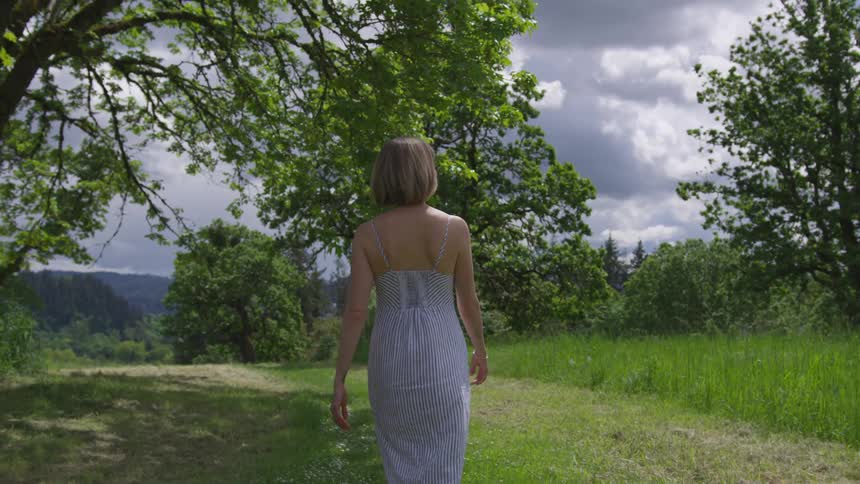 A woman wearing a striped sundress walks on a grassy trail on top of a hill near trees